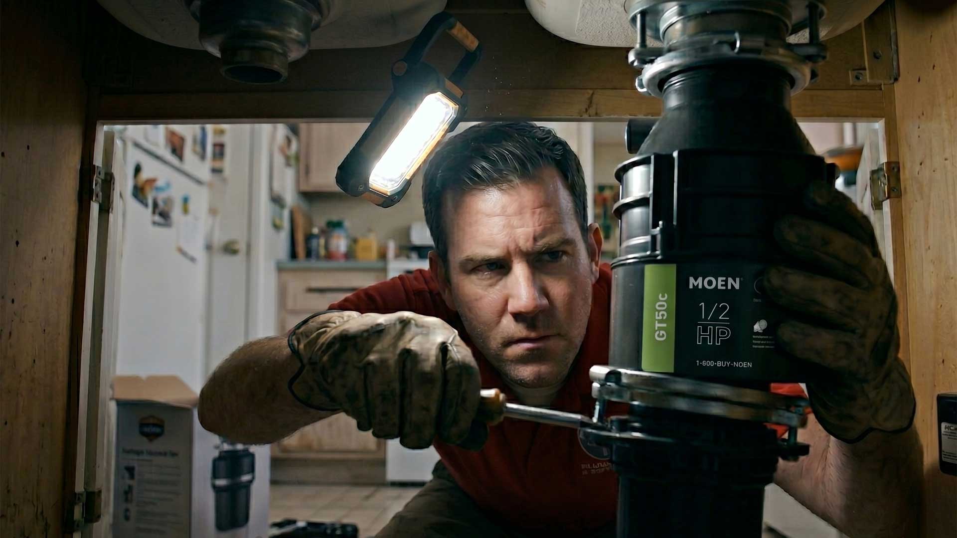 So-Cal 1 plumber mounting a new garbage disposal unit under a kitchen sink cabinet with a work light overhead